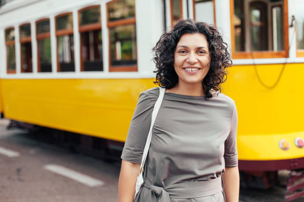 Uma mulher sorridente com cabelos cacheados e vestindo uma blusa cinza está de pé na frente de um bonde amarelo típico de Portugal. Ela exibe confiança e alegria, sugerindo que pode estar segurando seus Documentos de Identidade.